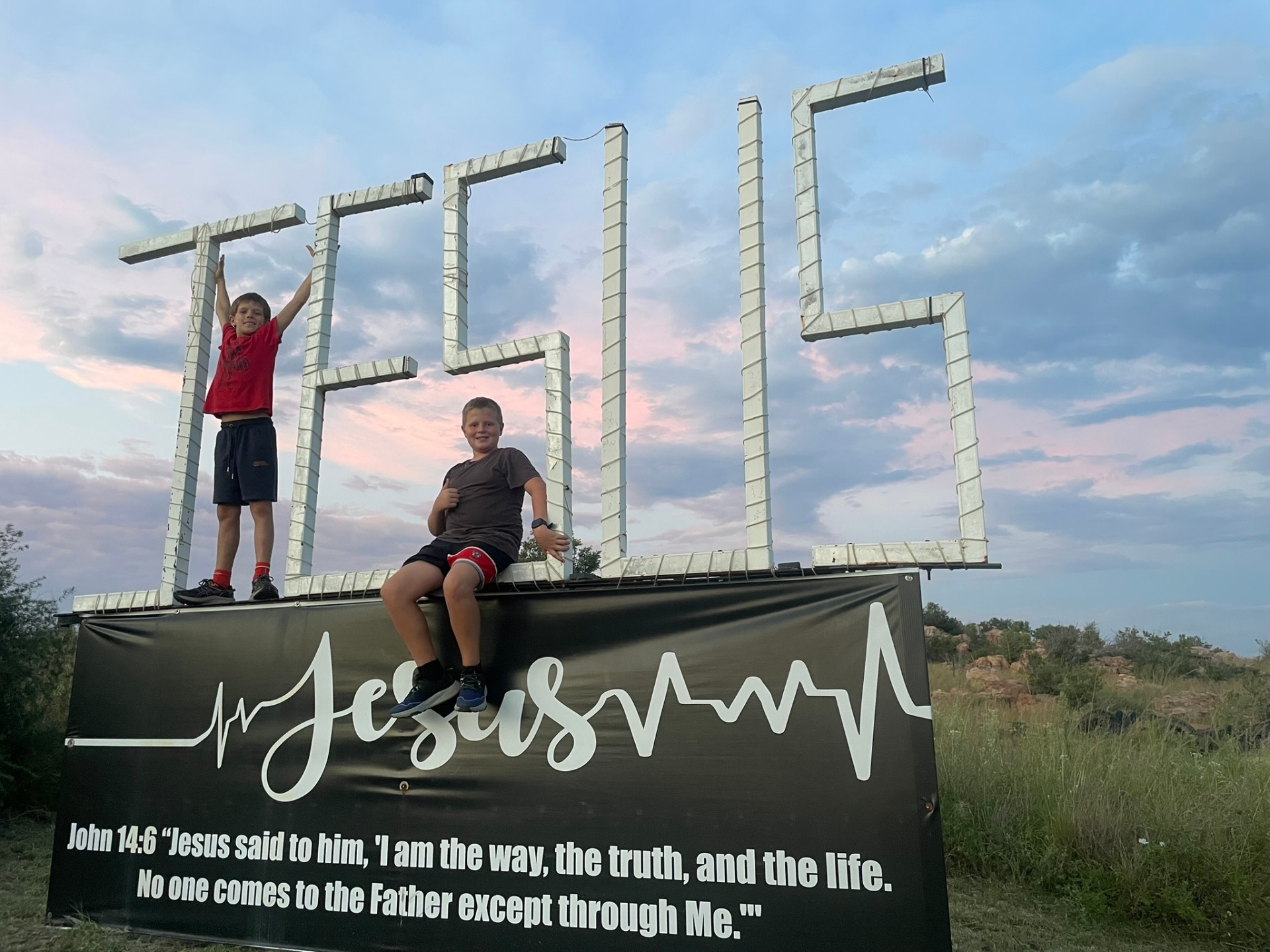 Two boys posing on top of the JESUS sign at dusk