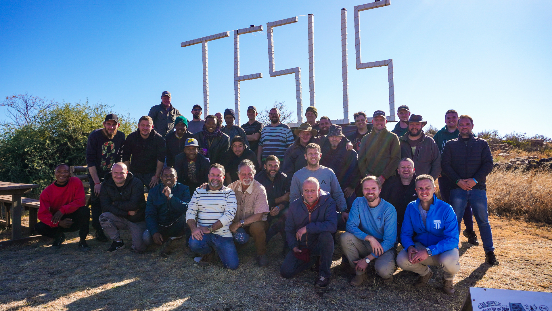 Men's group photo gathered beneath the JESUS sign on a clear blue-sky day