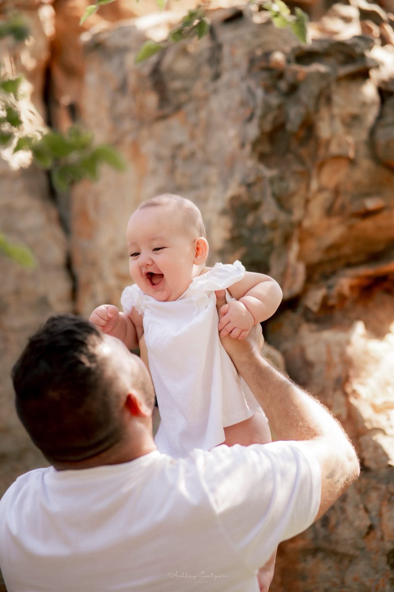 Dad lifting a giggling baby girl against a sunlit rock face